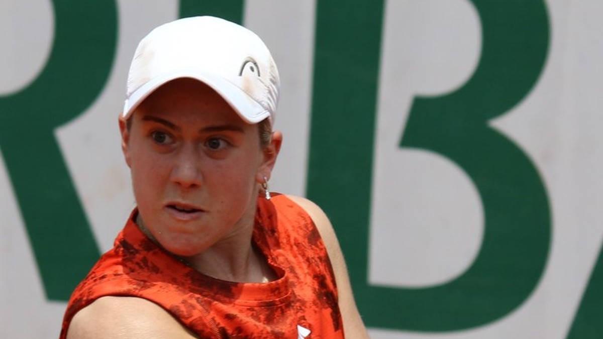 Sonay Kartal in a white cap and orange top follows through on a backhand in front of a green and white wall at the 2023 French Open