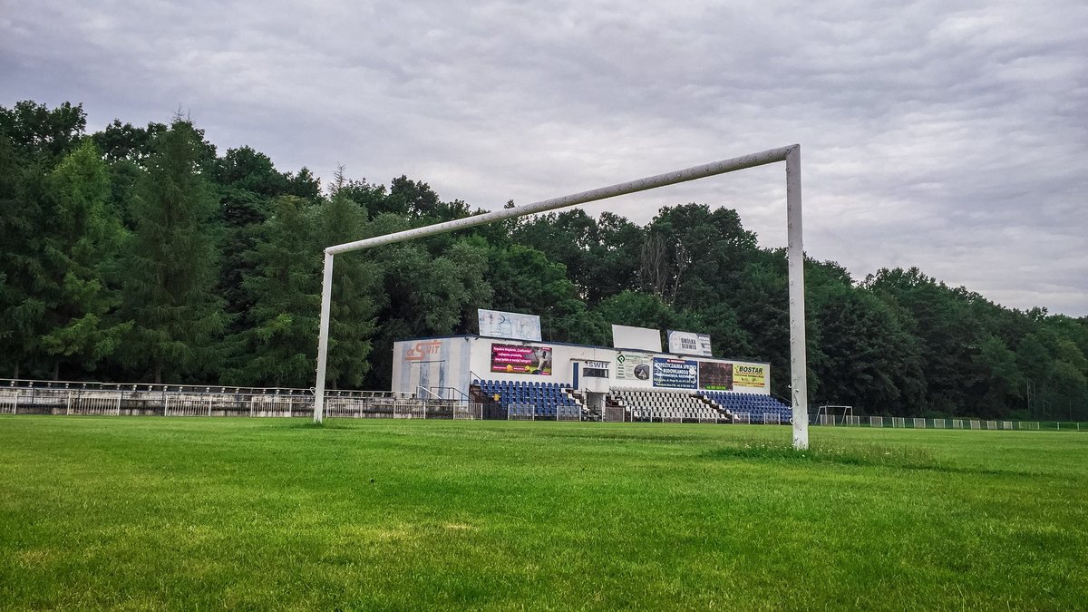 A soccer goal stands on an empty grass field in front of a small grandstand and trees