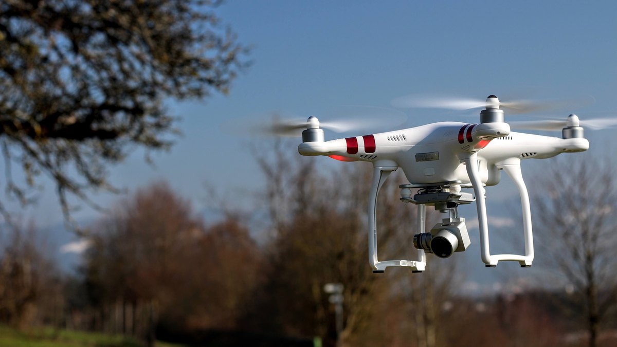 A white quadcopter drone hovering in the air above a park with trees behind it