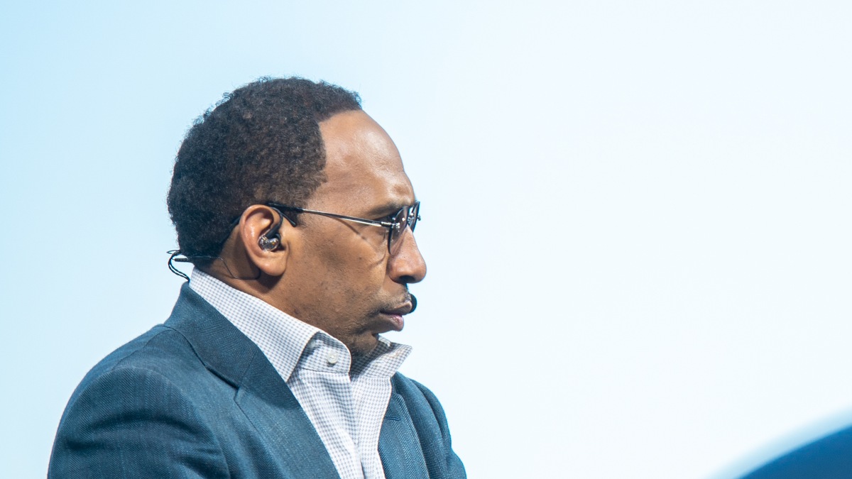 Stephen A. Smith in thin glasses and a blue suit jacket speaking in profile against a pale studio backdrop