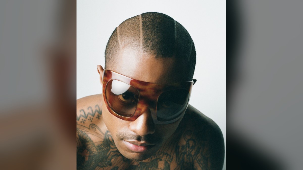Steve Lacy in oversized tortoiseshell sunglasses looking down at the camera against a pale studio backdrop