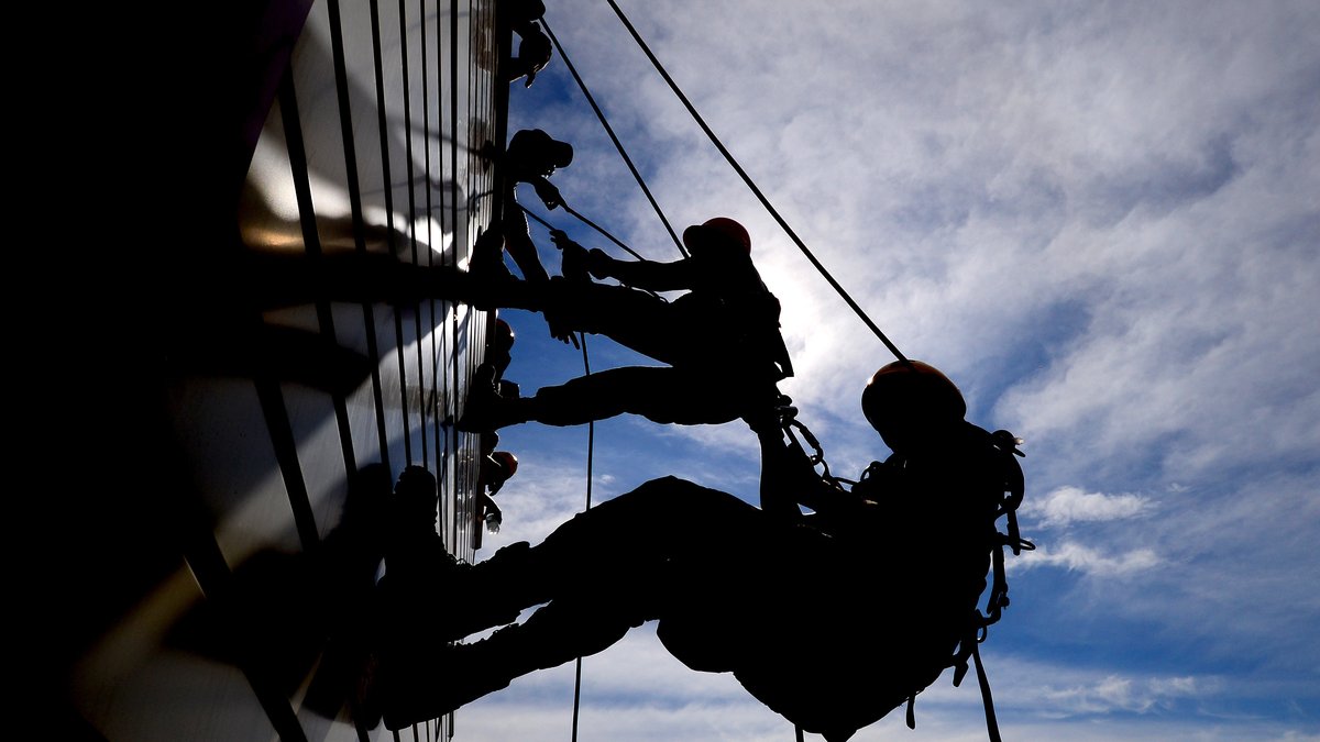 Silhouettes of two rescuers in helmets rappelling on ropes down the glass side of a tall building against a bright blue sky with clouds