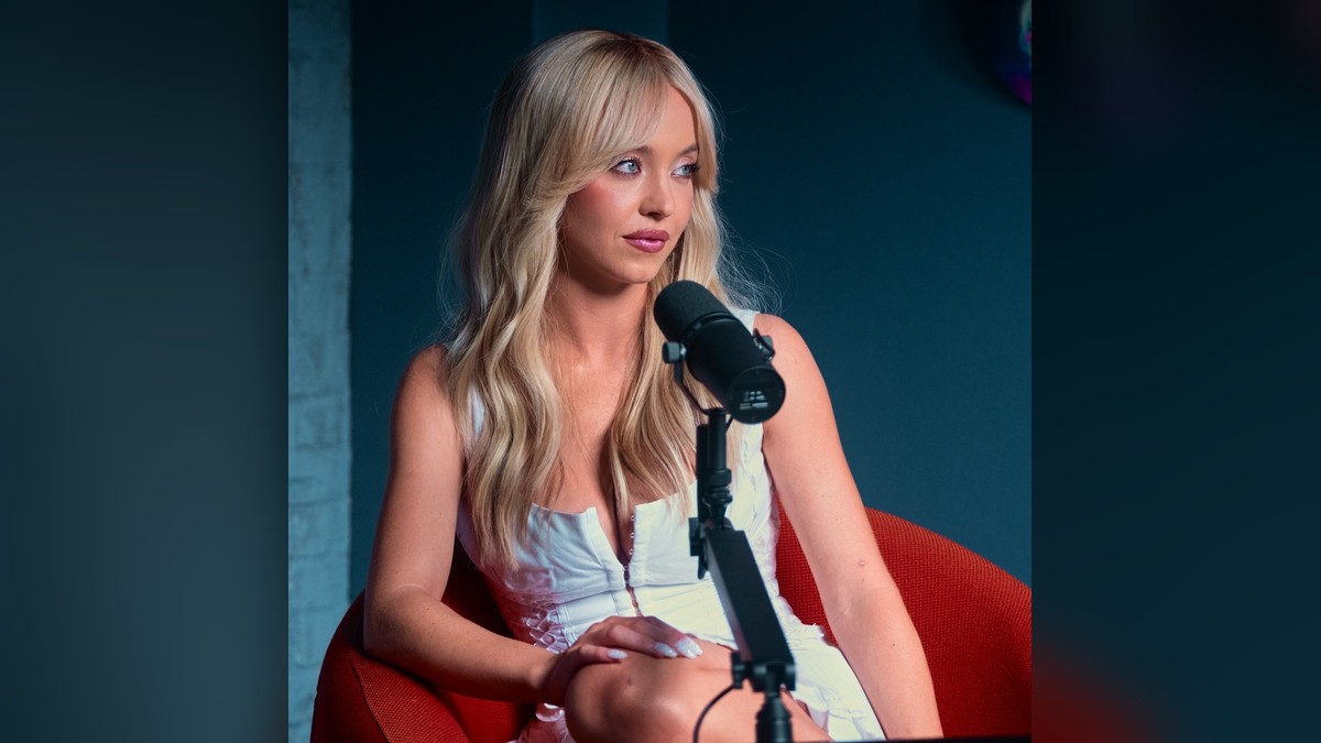 Sydney Sweeney in a white dress seated at a microphone on a red chair