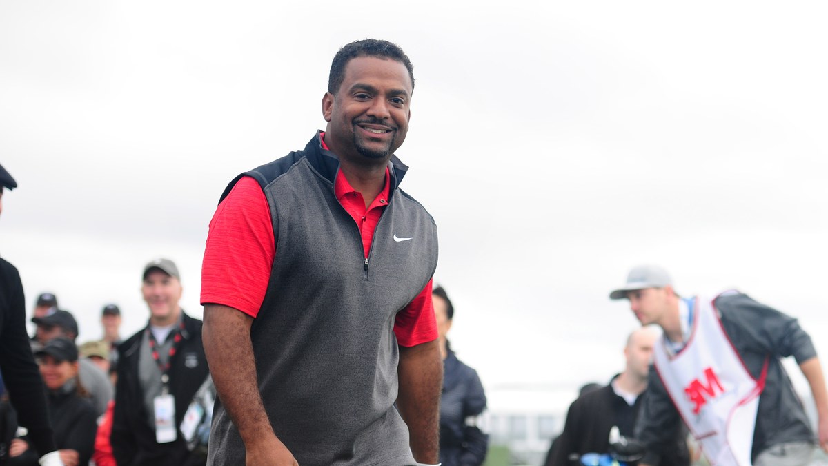 Alfonso Ribeiro grins in a red polo and gray vest as he walks across a golf course in front of a crowd