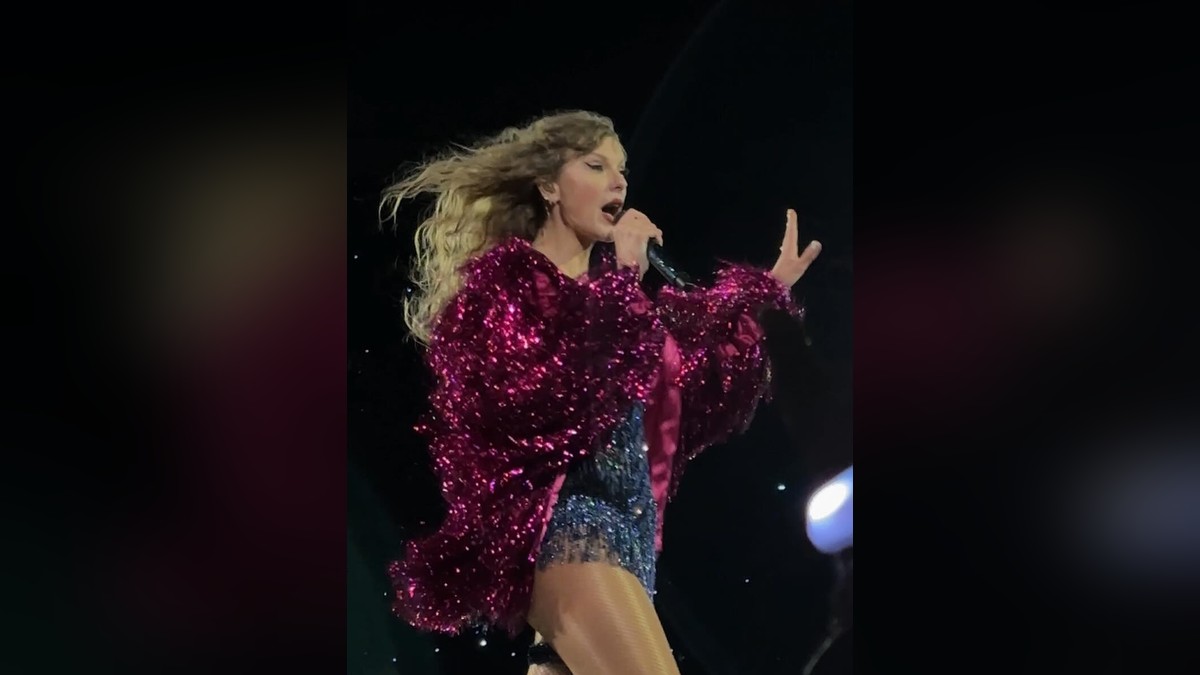 Taylor Swift singing into a microphone on stage in a sparkly pink fringed jacket