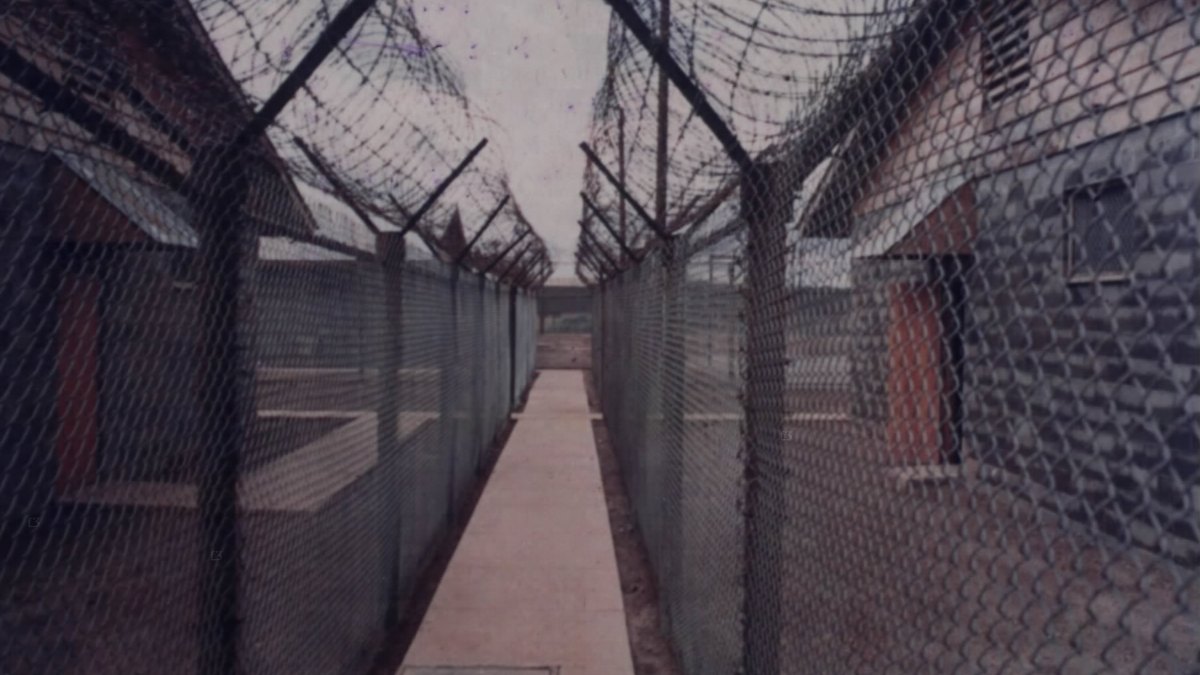 Tall chain-link prison fences topped with coiled razor wire lining a narrow walkway