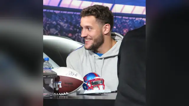 San Francisco 49ers defensive end Nick Bosa smiling in a gray hoodie at a table with football helmets during a Super Bowl fan event