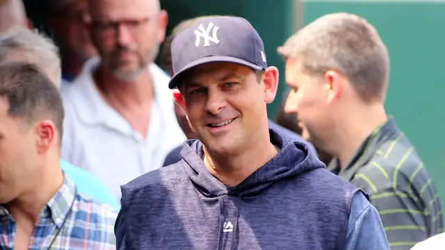 Aaron Boone smiles in a navy Yankees cap and sleeveless Yankees hoodie as he walks through a crowd in June 2018