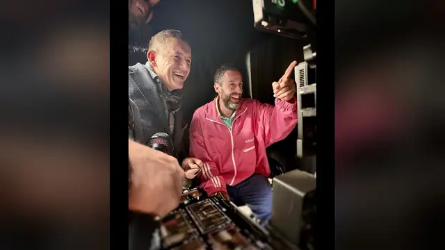 Adam Sandler in a pink jacket laughing and pointing beside Dany Boon on a film set