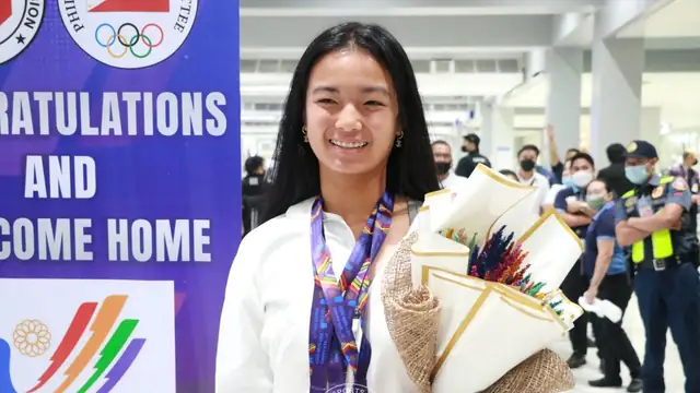 Alex Eala smiles in a white jacket with a medal lanyard around her neck while holding a bouquet of flowers on her arrival in Manila in 2022