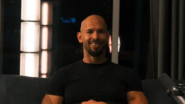 Andrew Tate smiling in a black T-shirt on a studio couch