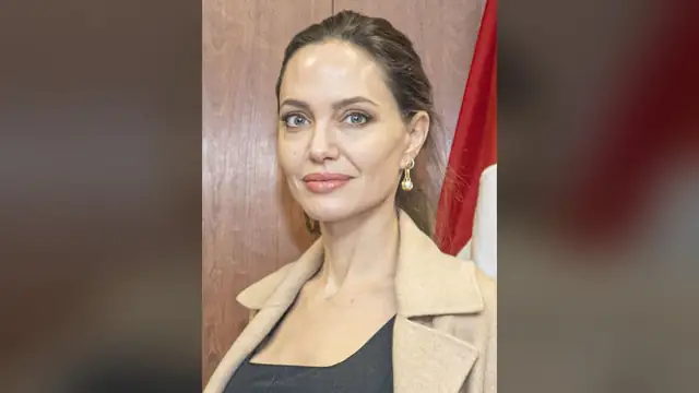 Angelina Jolie in a camel coat in front of a wood-paneled wall