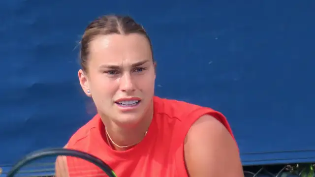 Aryna Sabalenka in a red sleeveless top grips her racket during a practice session in front of a blue wall