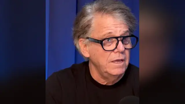 Anson Williams in black-framed glasses and a black T-shirt talking in front of a blue curtain