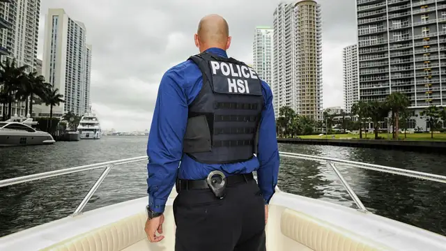A federal agent in a blue shirt and a black vest marked Police HSI stands on the bow of a boat facing city high-rises