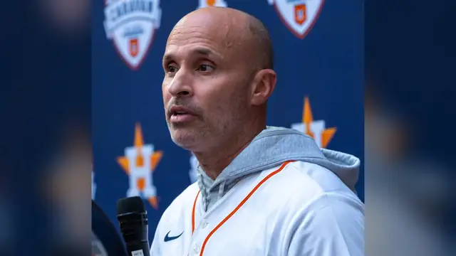 Houston Astros manager Joe Espada speaks into a microphone in a white Astros jersey and gray hoodie