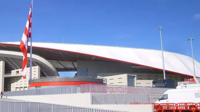 The curved white roof and red trim of Atlético Madrid's stadium under a clear blue sky with a red and white striped flag out front