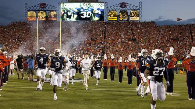 Auburn Tigers players in navy jerseys sprint onto the field at Jordan-Hare Stadium through smoke under the lights as fans pack the stands