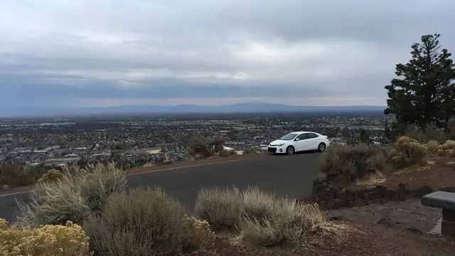 A white car parked on a road high above Bend, Oregon, under a gray cloudy sky