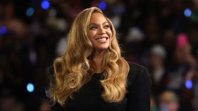 Beyoncé smiling with long honey-blonde waves in a black double-breasted blazer in front of a blurred crowd