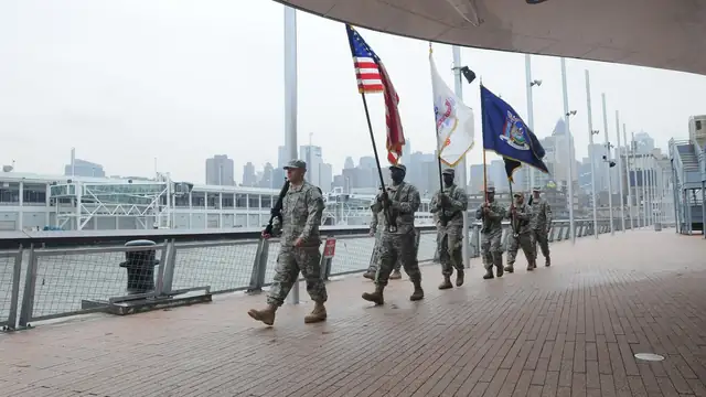 A military color guard carrying the American flag and unit flags marches across the Intrepid with the Hudson River and city towers behind them