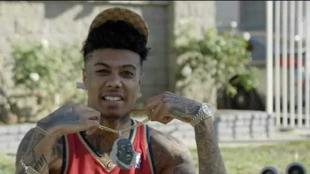 Blueface smiling in a red basketball jersey as he holds up a chain pendant outdoors