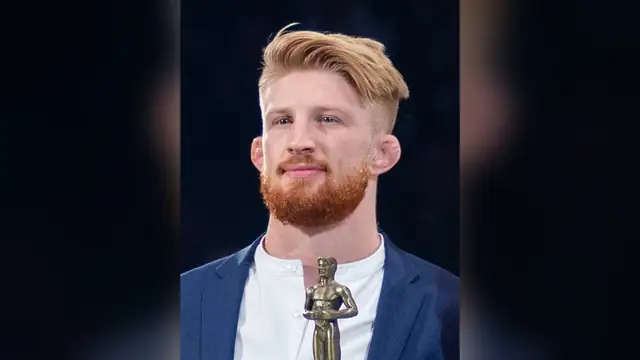 Bo Nickal in a navy blazer holds a small gold trophy during a Penn State wrestling ceremony