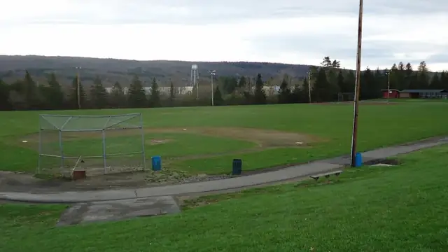 An empty baseball diamond with a dirt infield and green outfield beside a wooded hill