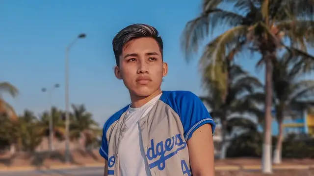 A young man in a gray and blue Dodgers baseball jersey standing on a sunny street lined with palm trees