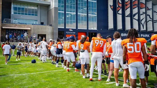 Denver Broncos players in orange jerseys stand on the practice field outside the Broncos Park team facility