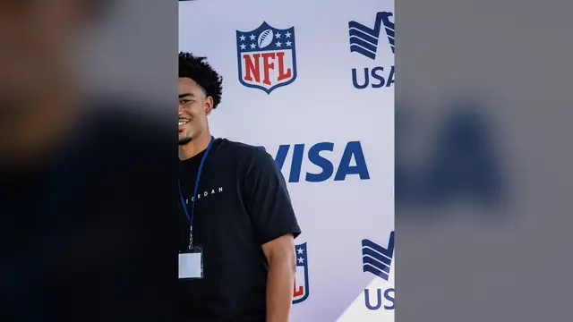 Bryce Young in a black T-shirt smiles in front of an NFL backdrop at a Fourth of July event at Camp Lejeune in 2024