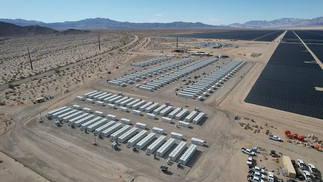 Rows of white battery storage containers stretch across a desert site beside a large solar farm