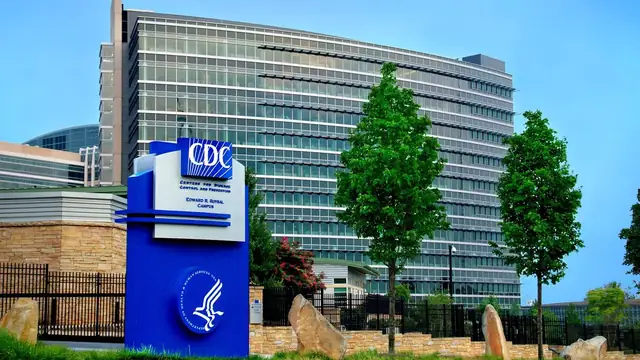 The blue CDC sign at the entrance to the agency's Roybal campus in Atlanta in front of a tall glass office tower and young trees