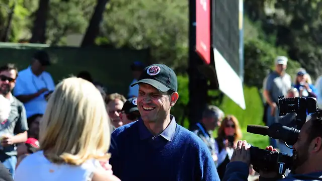 Jim Harbaugh smiles in a navy sweater and baseball cap as he walks through a crowd of fans and cameras at a Pebble Beach golf event