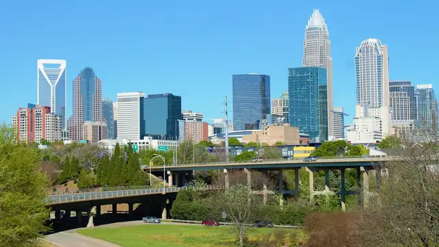 The Charlotte skyline rising beyond green trees and a highway bridge on a sunny day