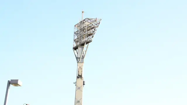 A stadium floodlight tower stands against a pale blue sky