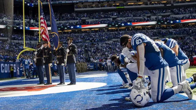 Indianapolis Colts players in blue and white uniforms kneel in a pregame prayer near the end zone as Army National Guard soldiers hold flags at Lucas Oil Stadium