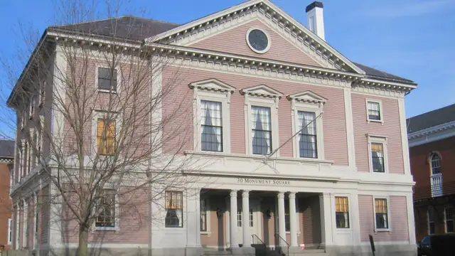 A historic brick building with tall white columns and a gabled roof facing Monument Square in Concord, Massachusetts under a blue sky