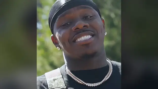 DaBaby smiling outdoors in a black cap and diamond chain
