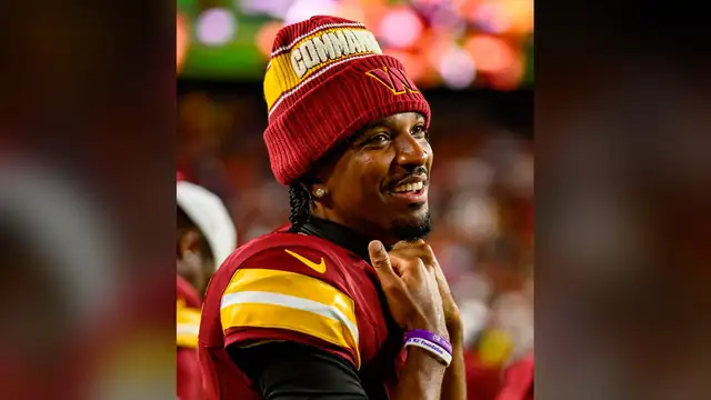 Commanders quarterback Jayden Daniels smiles on the sideline in a burgundy Commanders beanie and jersey during a home preseason game