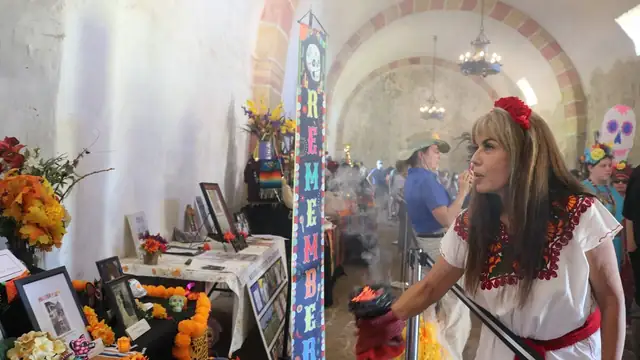 A woman in a white embroidered blouse and red flower in her hair stands beside Day of the Dead altars with marigolds inside a mission church