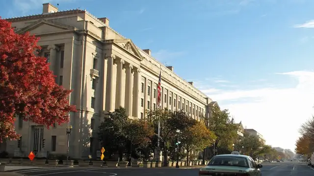 The limestone Robert F. Kennedy Department of Justice Building in Washington with an American flag out front, red autumn trees and a taxi on the wide avenue beside it