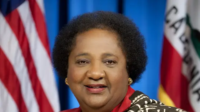 California Secretary of State Shirley Weber smiling in a red blouse and patterned jacket between the American and California flags