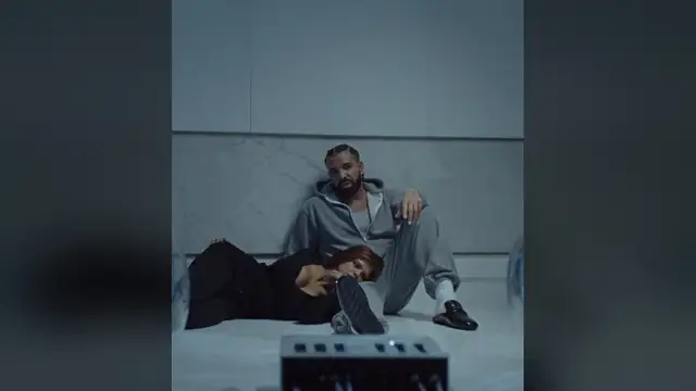 Drake in a gray hoodie sitting on a studio floor beside a woman in a black top during the Janice STFU video