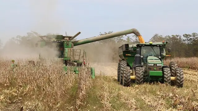 A combine unloads harvested corn into a grain cart pulled by a green tractor