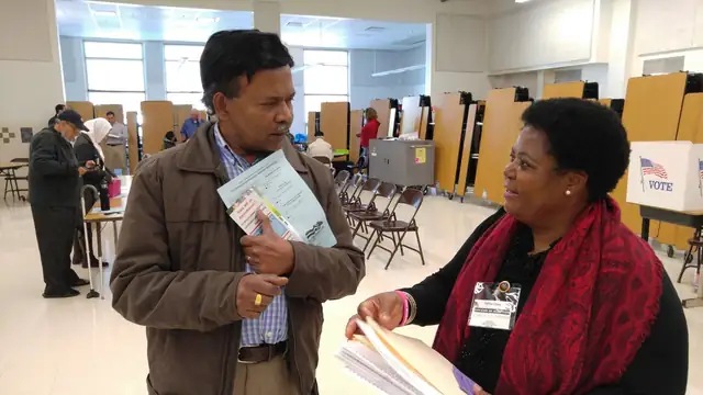 A voter holding a sample ballot talks with an election officer in a red scarf inside a school polling place lined with voting booths