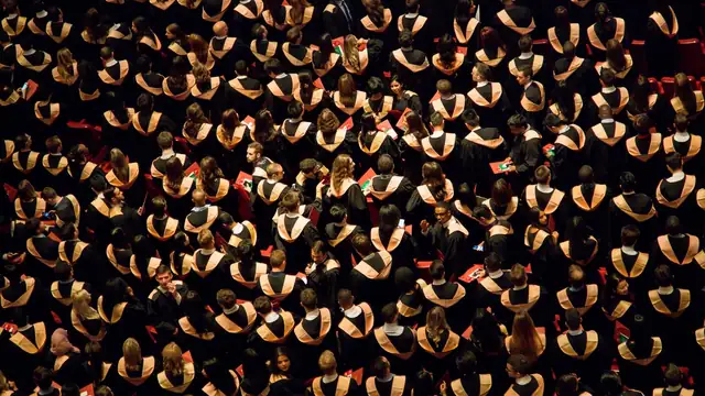 Hundreds of graduates in black caps and gowns seen from above filling an outdoor ceremony