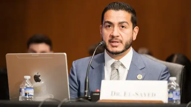Abdul El-Sayed in a blue suit speaks into a microphone at a witness table with a nameplate in front of him and a wood paneled wall behind