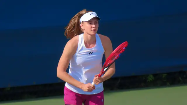 Elena Rybakina in a white top and visor holding her red racket on a hard court