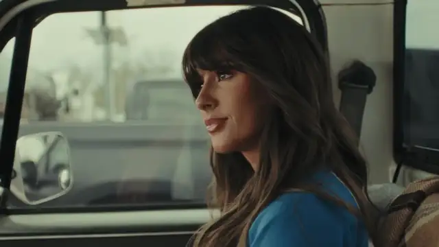 Ella Langley with long brown hair and bangs smiling softly in a blue top inside a truck